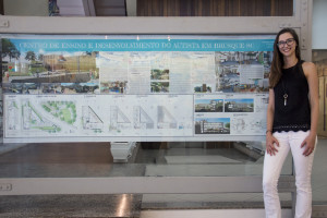 Fotografia colorida em ambiente fechado. Hall da reitoria. A estudante Manoela Fischer posa ao lado da prancha com o projeto arquitetônico do seu TCC, em que se lê, no topo, “Centro de Ensino e Desenvolvimento do Autista em Brusque-SC”.